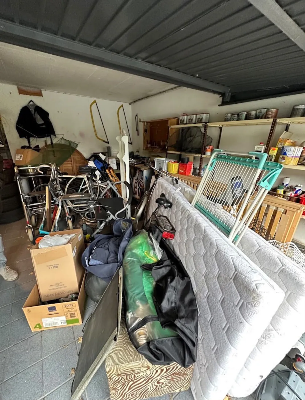 Vollgestellte Garage mit Matratzen, Fahrrädern, Kartons und Regalen vor der Entrümpelung